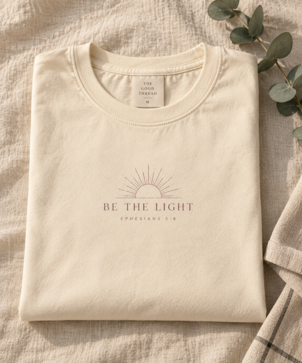 Butter Cream relaxed-fit tee printed with “Be the Light” in mauve serif type, folded on a linen backdrop