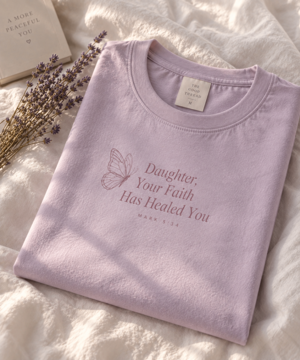 Lilac relaxed-fit tee printed with “Daughter, Your Faith Has Healed You” in mauve serif type, folded on a linen backdrop