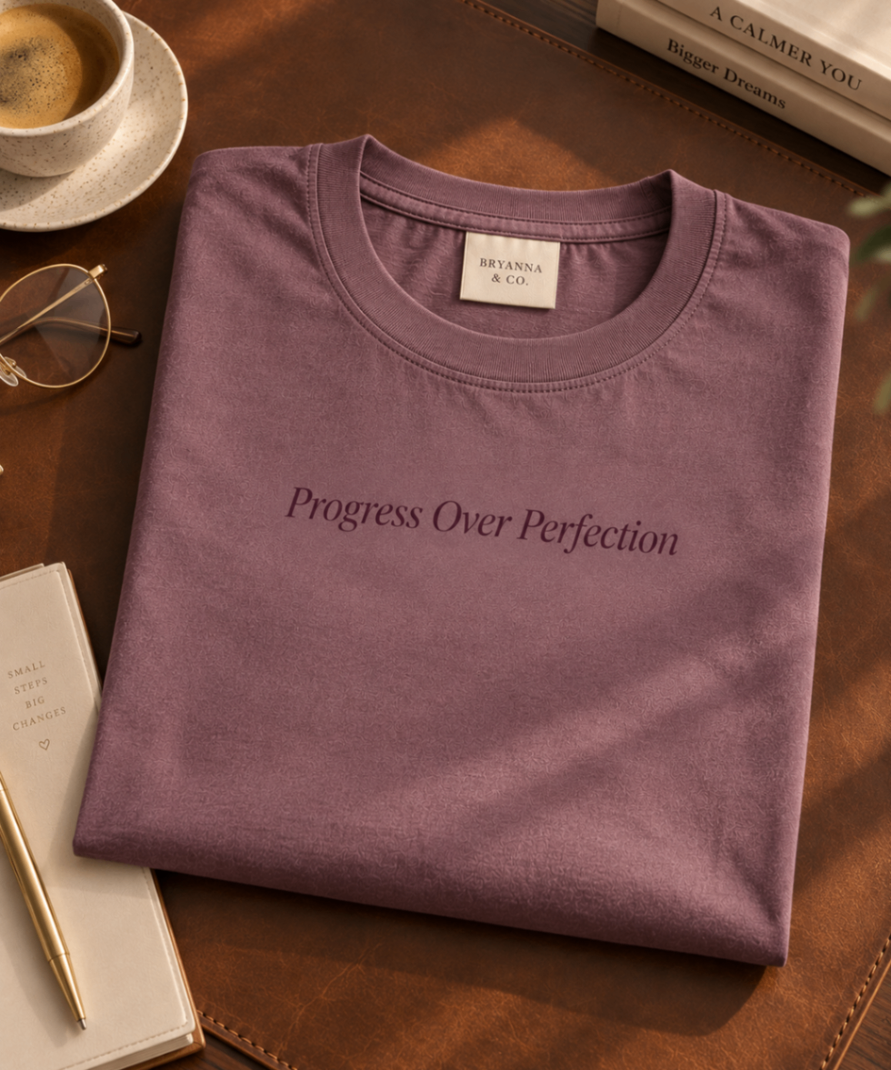 Soft Plum relaxed-fit tee printed with “Progress Over Perfection” in mauve serif type, folded on a linen backdrop