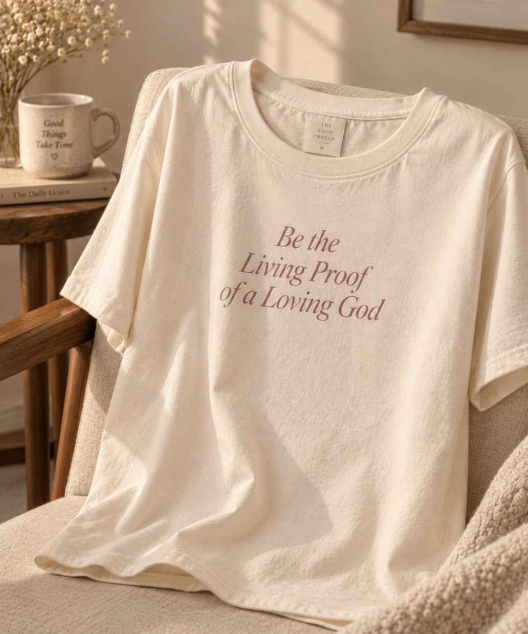 Warm Ivory relaxed-fit tee printed with “Be the Living Proof of a Loving God” in mauve serif type, folded on a linen backdrop