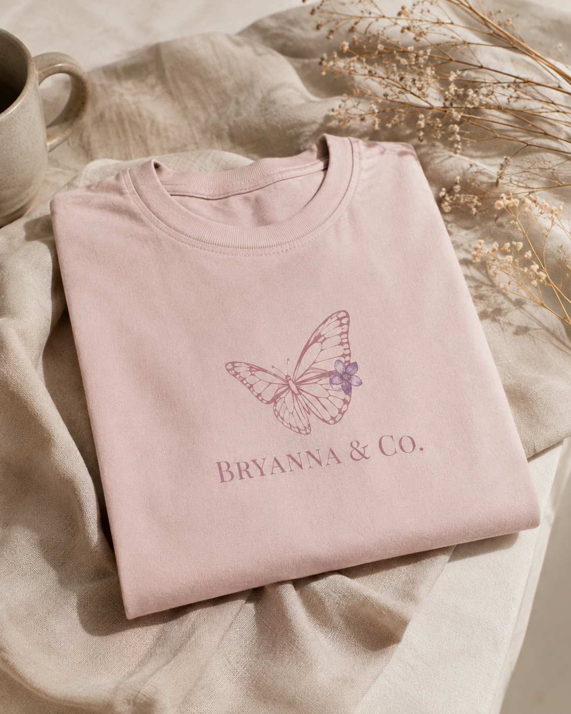 A folded blush tee on a linen backdrop beside a ceramic mug and dried gypsophila