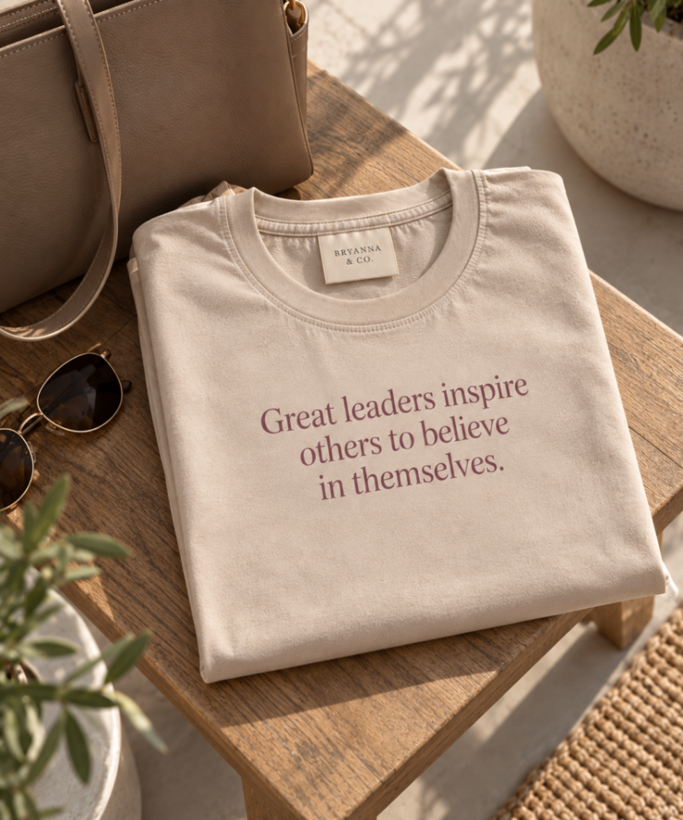 Warm Sand relaxed-fit tee printed with “Great leaders inspire others to believe in themselves.” in mauve serif type, folded on a linen backdrop