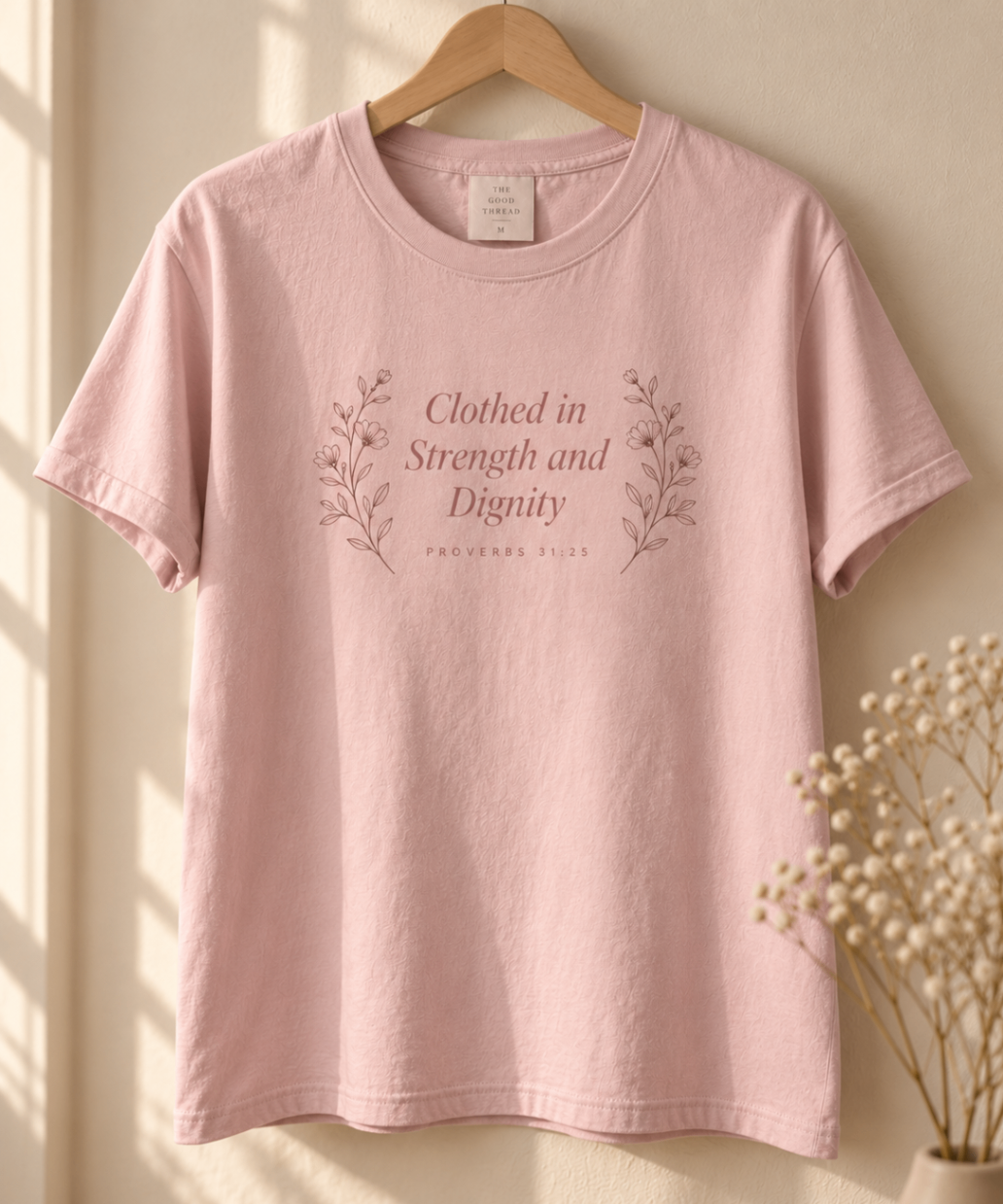 Powder Pink relaxed-fit tee printed with “Clothed in Strength and Dignity” in mauve serif type, folded on a linen backdrop