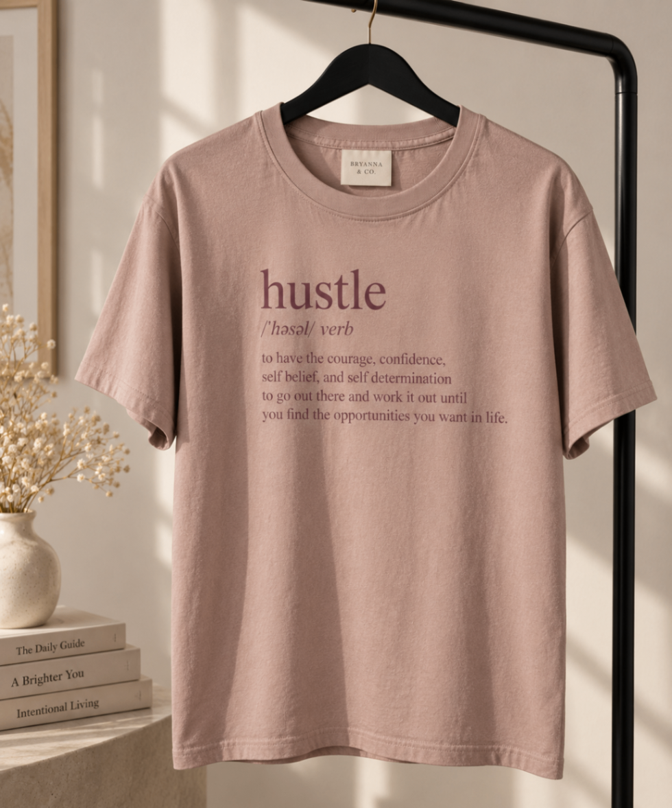 Taupe Pink relaxed-fit tee printed with “hustle · /ˈhəsəl/ verb · to have the courage, confidence, self belief, and self determination to go out there and work it out until you find the opportunities you want in life.” in mauve serif type, folded on a linen backdrop