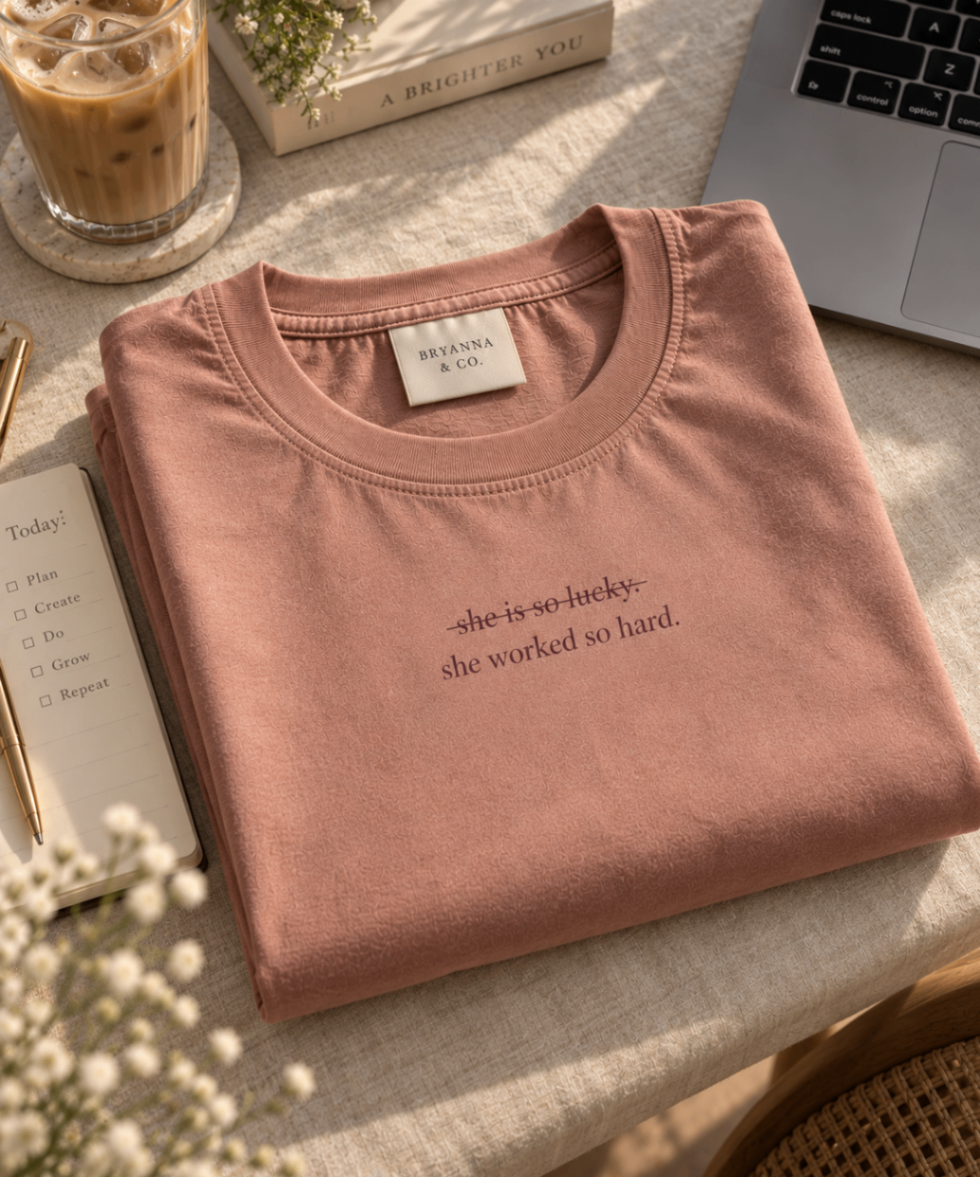 Terracotta Pink relaxed-fit tee printed with “she is so lucky. she worked so hard.” in mauve serif type, folded on a linen backdrop